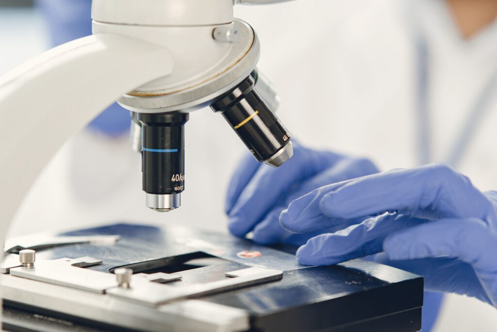 Lab technician in blue gloves examining a sample under a microscope, representing quality-control testing at a Malaysian medical-grade lubricant and latex manufacturer.