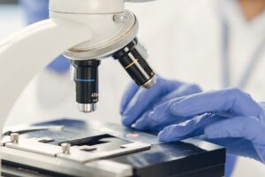 Lab technician in blue gloves examining a sample under a microscope, representing quality-control testing at a Malaysian medical-grade lubricant and latex manufacturer.