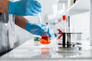 Gloved lab technician testing an orange-tinted formulation in a glass flask, representing SPF sunscreen formulation quality testing at a Mexican cosmetics OEM manufacturer.